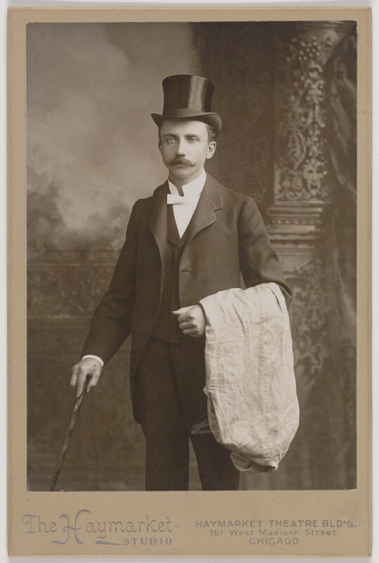 A black-and-white studio photograph of a White man with a mustache, wearing a top hat and three-piece suit, holding a cane in one hand, white cloth draped over the other arm.