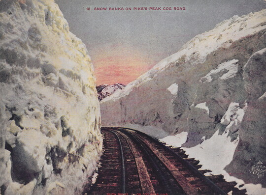 A color postcard photograph of railroad tracks curving through tall walls of snow on either side; a mountain peeking over the snow in the distance.