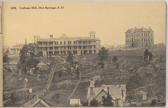 A sepia-toned photograph of, in the foreground, a three-story house with wide verandas on each level on a hill and several multi-story buildings in the background.