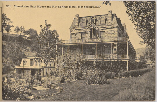 A sepia-toned postcard of a three-story stone hotel with a wrap-around porch nestled in a lush landscape.