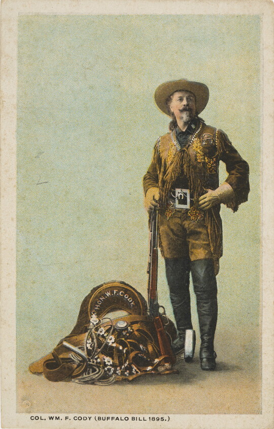 A color photograph postcard of a White man wearing a cowboy hat, brown leather jacket with fringe, tall black boots over his pants posing with a rifle and a pile of equestrian equipment.