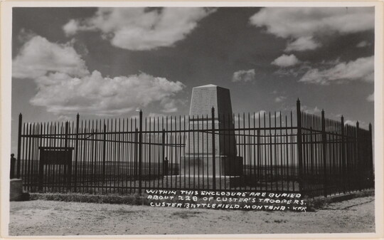 A black-and-white photograph postcard of a stone monument surrounded by a black iron fence beneath a cloudy sky.