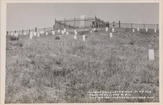 A black-and-white photograph postcard of a graveyard on a hillside below a large central monument with a fence around it.