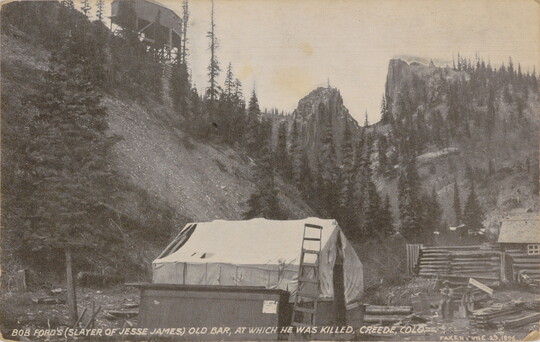 A sepia-toned postcard of a small building with a white tarp as the roof at the foot of steep mountains.