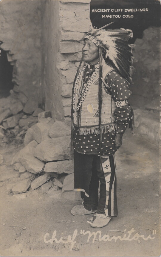 A black-and-white photograph of a Indigenous man in ceremonial dress standing in front of stone dwelling.