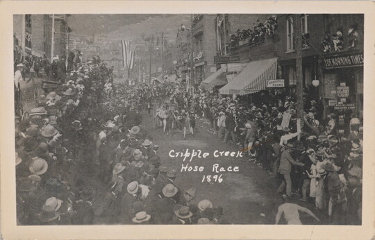 A black-and-white postcard of crowds of people lining a town street watching people harnessed to carriages running.