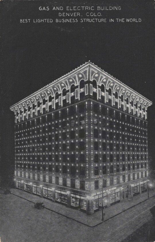 A color postcard of a nine-story building lit up at night under a full moon that is partially hidden behind clouds.