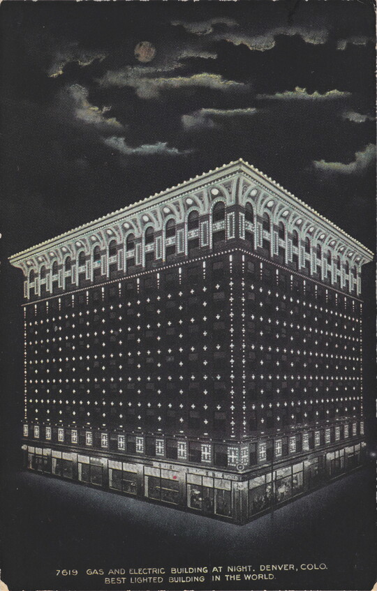 A color postcard of a nine-story building lit up at night under a full moon that is partially hidden behind clouds.