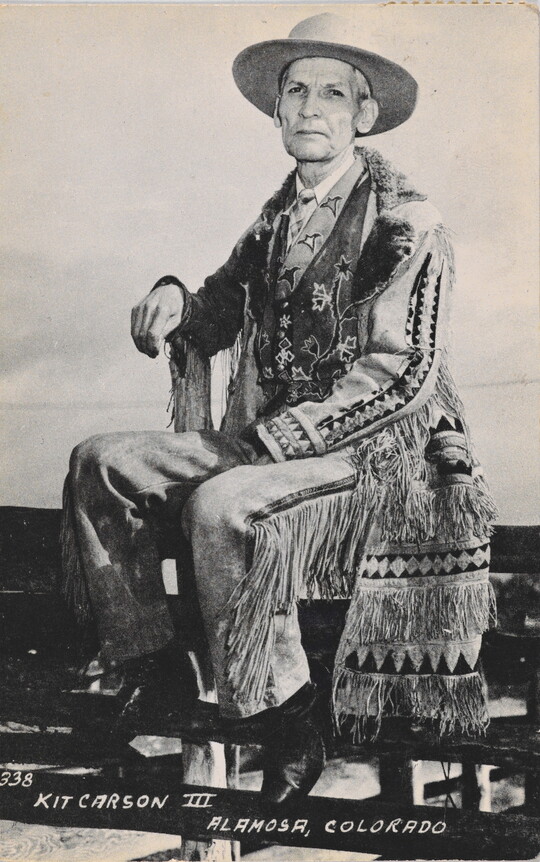 A black-and-white postcard of an older White man in western dress seated on a fence.