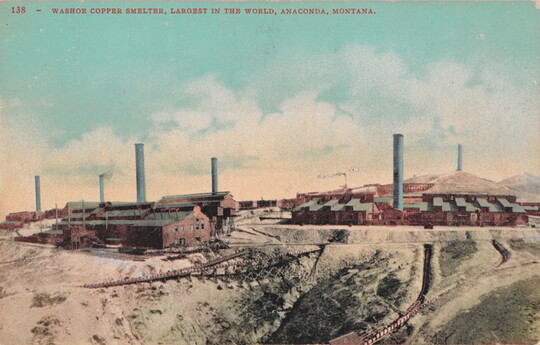 A color postcard of several industrial brick buildings with smokestacks in a mostly dirt terrain.