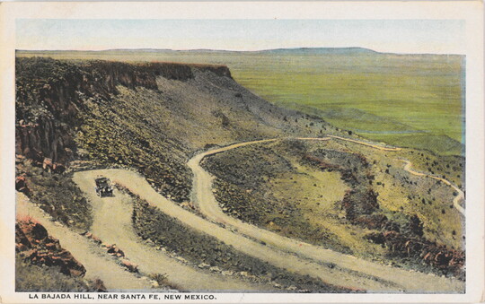 A color postcard of a high mesa with a car driving on a road that zig-zags back and forth down its side.