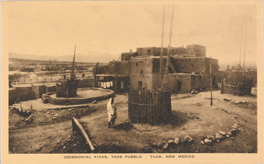 A sepia-toned photograph on a postcard of an adobe pueblo next to several kivas.