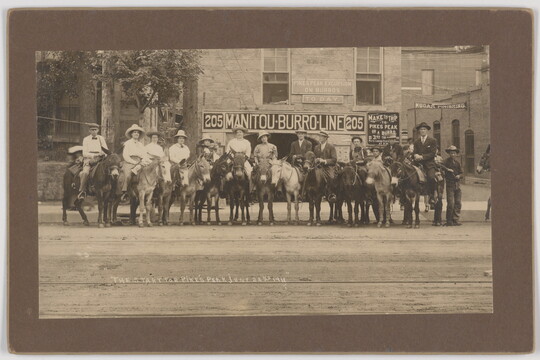 A sepia-toned photograph of a group of people, most with cowboy hats, on horses or donkeys on a dirt road in front of a brick building.