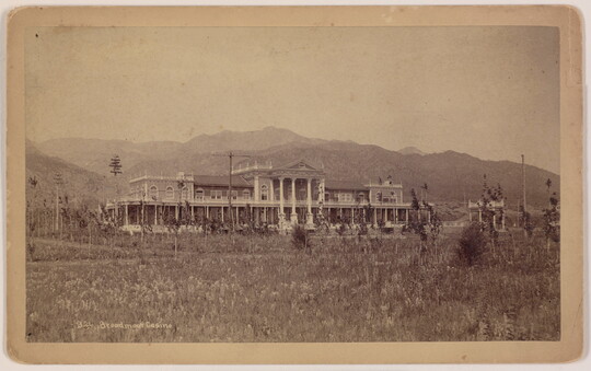 A sepia-toned photograph of a large, multi-story ornate building and mountains in the background.