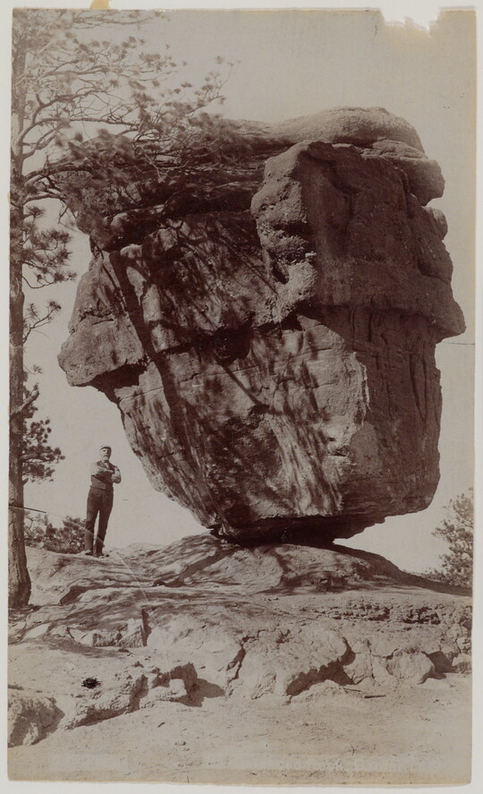 A black-and-white photograph of a person standing next to a large rock formation that is balancing on a narrow end.