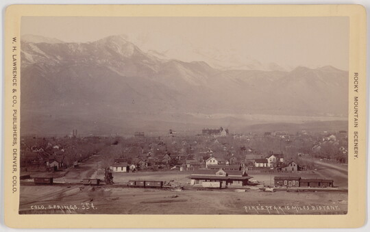 A black-and-white photograph of a railroad and station in the foreground, then a town, and snow-covered mountains in the background.