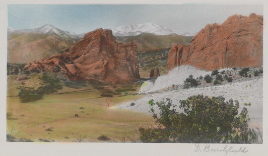 A color photograph of a plain at the foot of tall, red rock formations and snowy mountains in the distance.