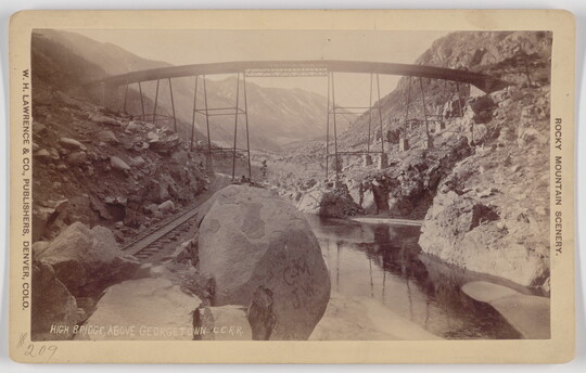 A black-and-white photograph postcard of a very tall simple truss bridge spanning railroad tracks and a deep rocky valley and river.