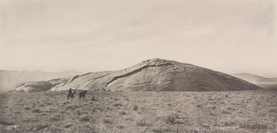 A black-and-white photograph of two people and two horses in a grassy field in front of a very large, smooth rock that seems to be emerging from the ground.