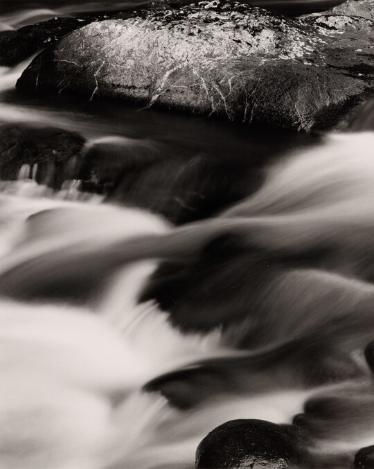 A black-and-white photograph of a water rushing over dark rocks.