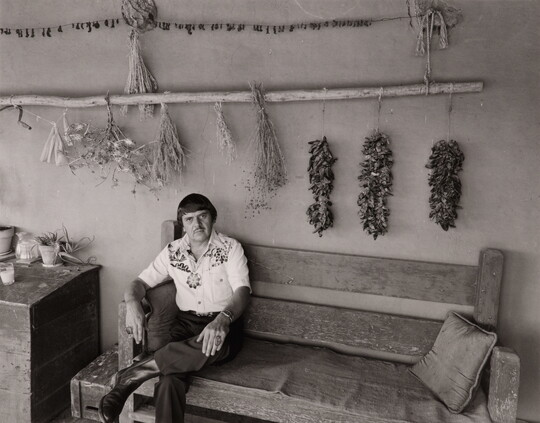 A black-and-white photograph of a man sitting on a bench under a long pole that has dried chili peppers and other herbs hanging from it.