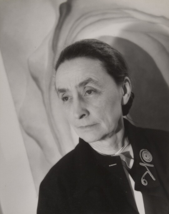 A black-and-white portrait photograph of an older White woman in front of a painting.