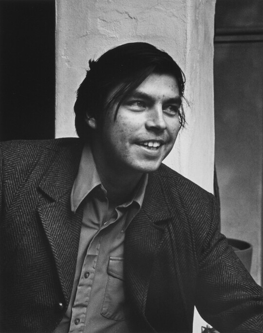 A black-and-white portrait photograph of a young medium-skinned man with longish dark hair, smiling, wearing a jacket over a button-down shirt.