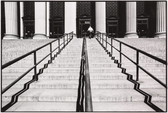 A black-and-white photograph of a person walking up a large set of stairs lined with handrails toward a building with large fluted columns.
