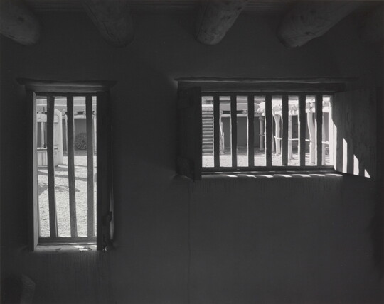 A black-and-white photograph taken from inside a dark room with a low ceiling looking toward a door and window with bars in them.
