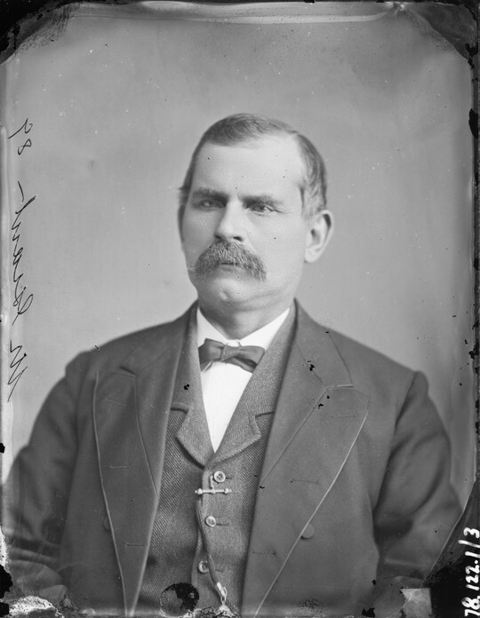 A black-and-white portrait image of a balding White man with a moustache wearing a dark three-piece suit with a bow tie.