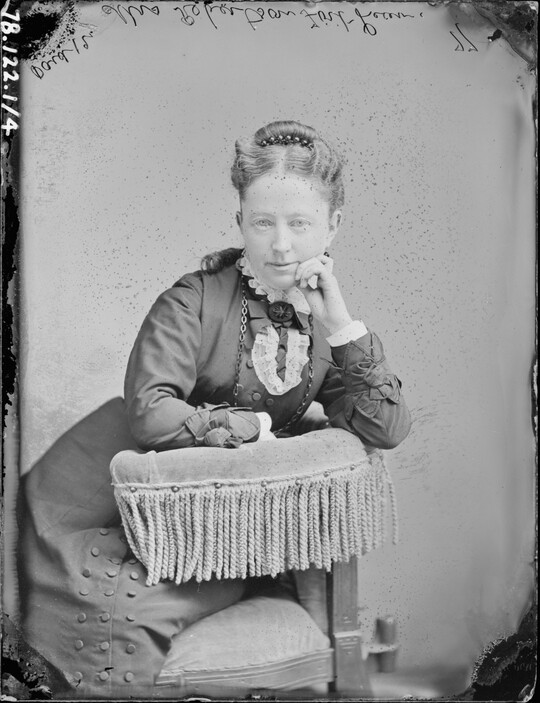 A black-and-white portrait image of a seated White woman, hair pulled to the top of her head, wearing a high-collared dress, leaning on the fringed arm of a chair, one hand raised to her cheek.