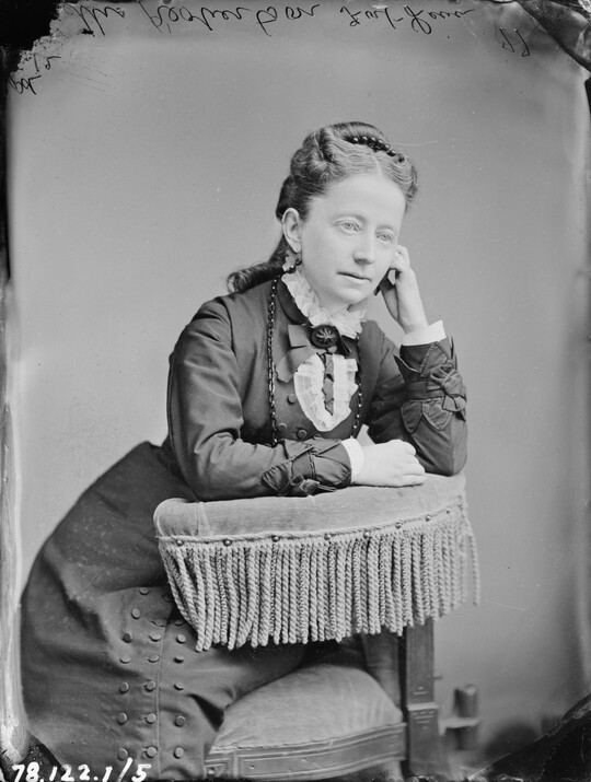 A black-and-white portrait image of a seated White woman, hair pulled to the top of her head, wearing a high-collared dress, leaning on the fringed arm of a chair, one hand raised to her cheek.