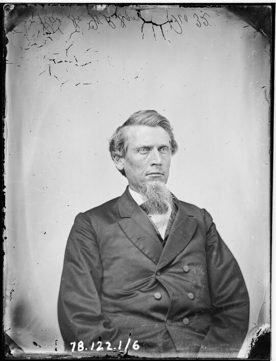 A black-and-white portrait image of a seated White man with a long goatee wearing a double-breasted jacket.
