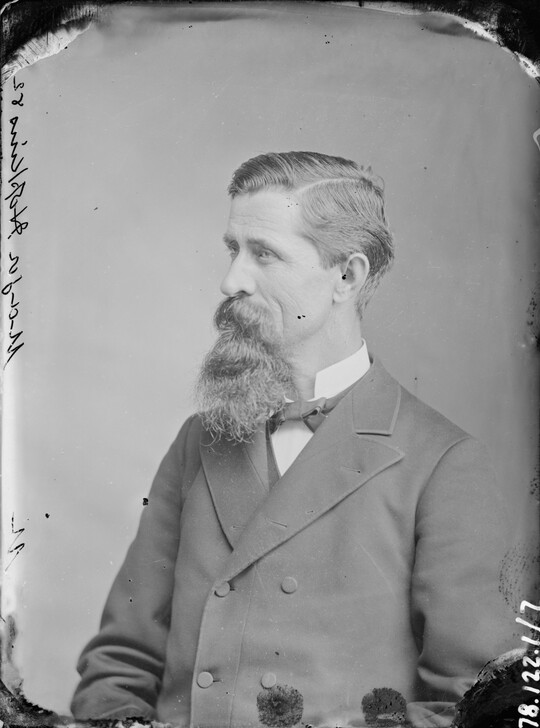 A black-and-white portrait image of a seated White man with a long mustache and goatee wearing a bowtie and double-breasted jacket.