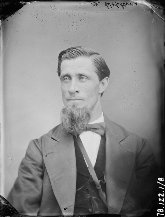 A black-and-white portrait image of a seated White man with a long goatee wearing a bowtie, vest, and jacket.