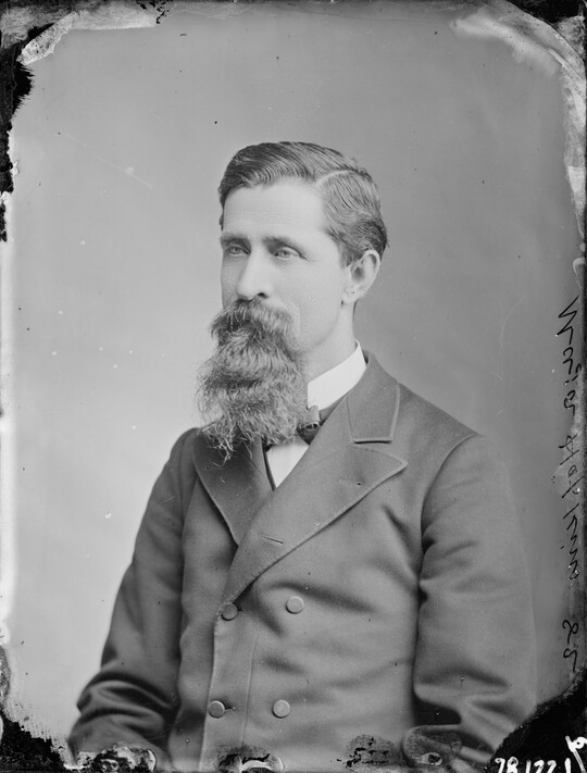 A black-and-white portrait image of a seated White man with a long mustache and goatee wearing a bowtie and double-breasted jacket.