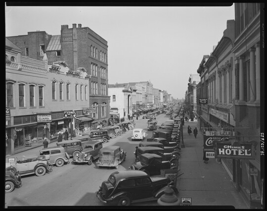 A black-and-white image of a street lined with tall buildings and parked automobiles.