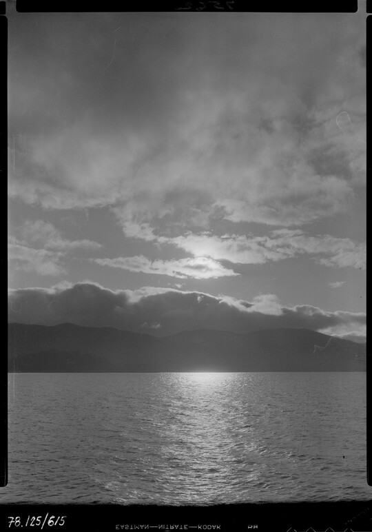 A black-and-white image of a lake, mountains on the opposite shore, under a cloudy sky at sunset.