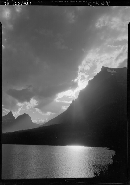 A black-and-white image of a cloudy sky with the sun setting behind a mountain overlooking a still lake.