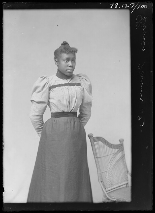 A black-and-white portrait image of a formally-dressed young Black woman, hair pulled up on her head, standing next to a wicker chair.