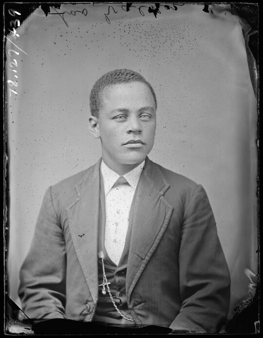 A black-and-white studio image of a seated young Black man with short dark hair, wearing a three-piece suit with a watch chain dangling from the vest.