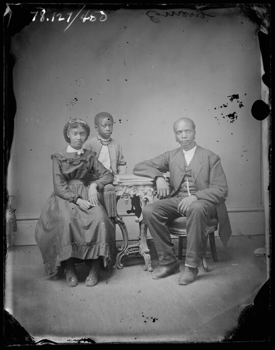 A black-and-white portrait image of a Black family that shows a man and young woman sitting at a table and a child standing behind the woman.