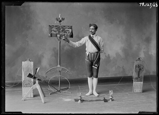 A black-and-white image of a man standing in front of a sign that reads, "The Great Layton," and other performance props.
