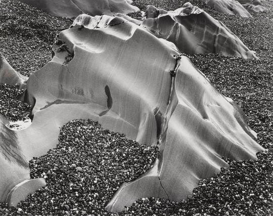 A black-and-white photograph of smooth, large, weather-worn rocks poking above a beach of smaller pebbles.