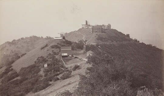 A sepia-toned photograph of a domed building and other structures atop a steep hillside covered with vegetation.