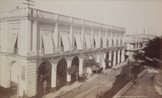 A black-and-white photograph of a white two-story hotel with arched entrances to an arcade on the street level.