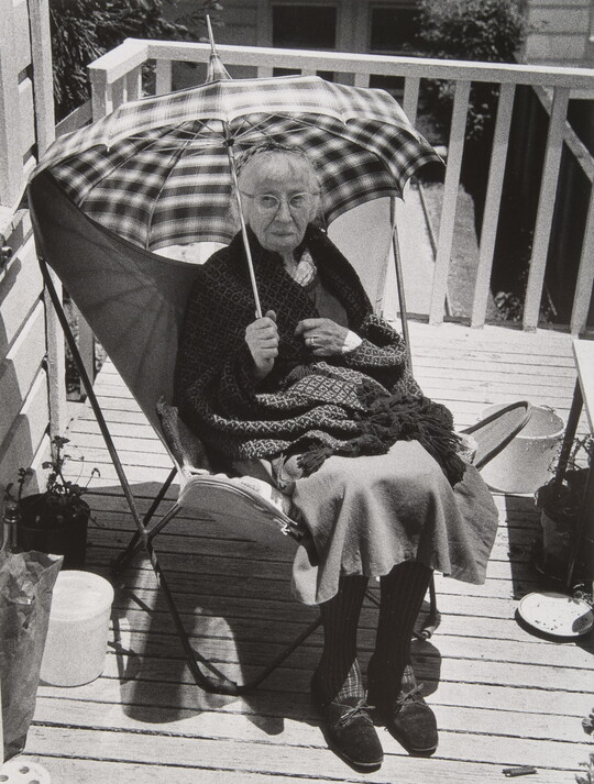 A black-and-white photograph of an older White woman wearing glasses, a blanket on her lap, holding an umbrella while sitting in a chair on a wood deck.
