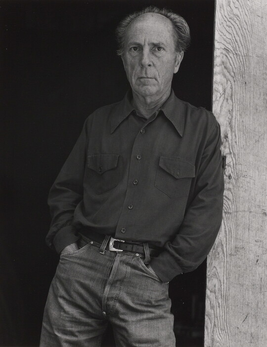 A black-and-white portrait of an older, balding White man wearing jeans and a long-sleeve button-down shirt leaning against a wooden beam.