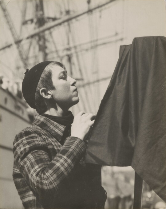A black-and-white portrait photograph of a young, White woman in profile with short hair, wearing a beanie, standing behind a camera covered in black cloth.