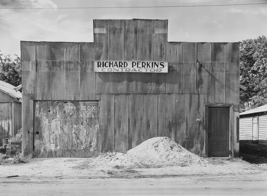A black-and-white photograph of a building with a boarded up window and a pile of sand or gravel in front.